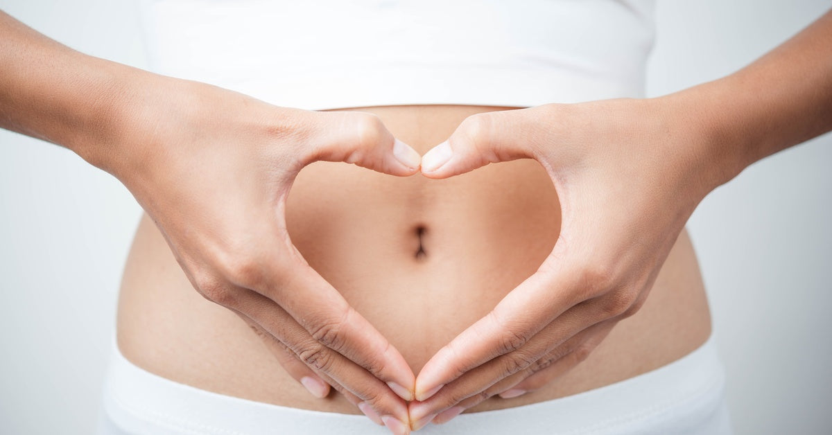 A close-up of a woman's stomach. She's making a heart symbol with her hands over her belly button.