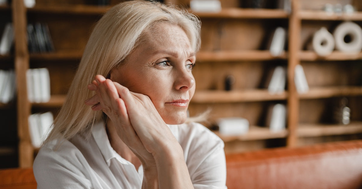 A woman sits on a couch and has her hands wrapped up holding her face up as she solemnly looks to the side.