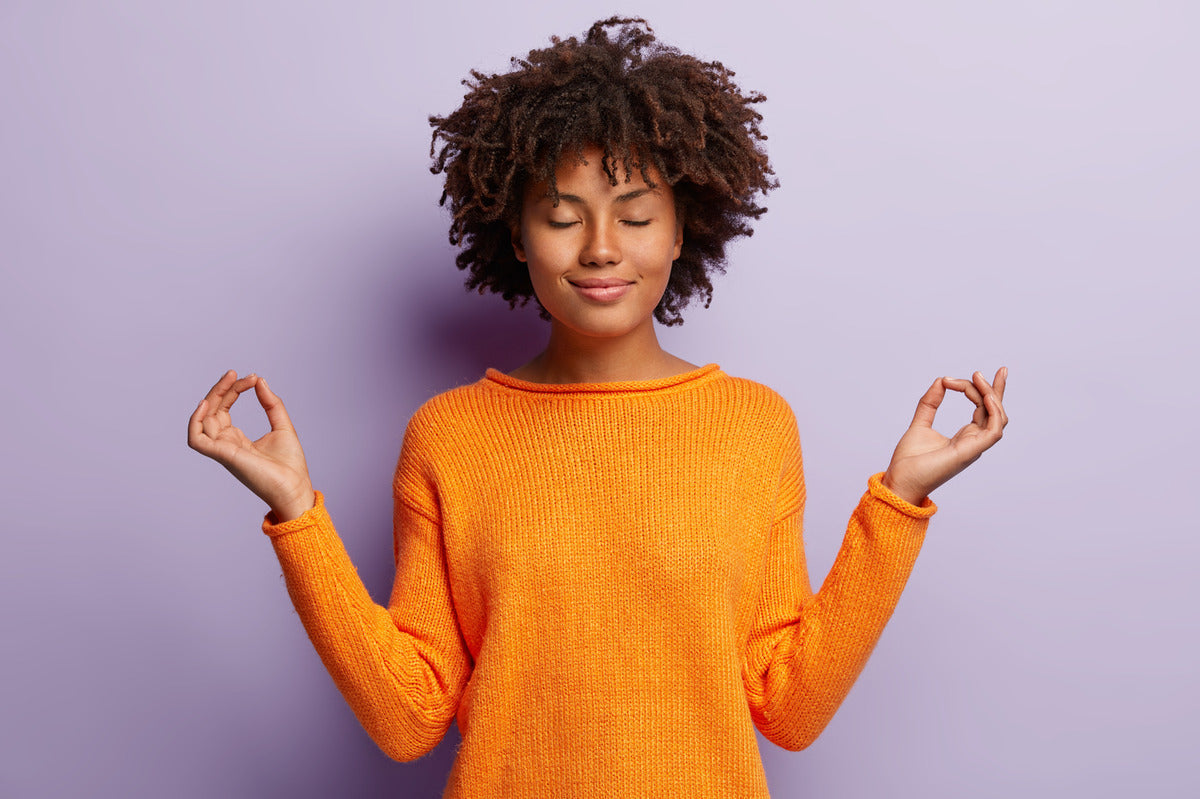 A woman standing in front of a purple wall with her eyes closed. She touches her forefingers to her thumbs.