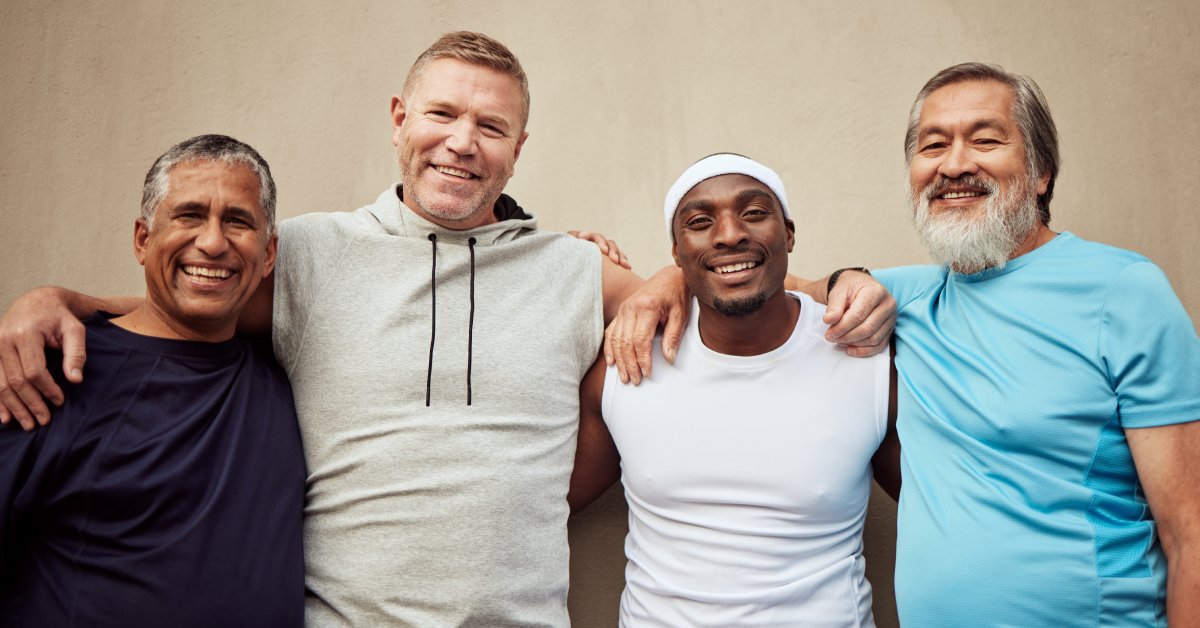 A group of men stand together with their arms around each other, smiling. The wall is bare behind them.