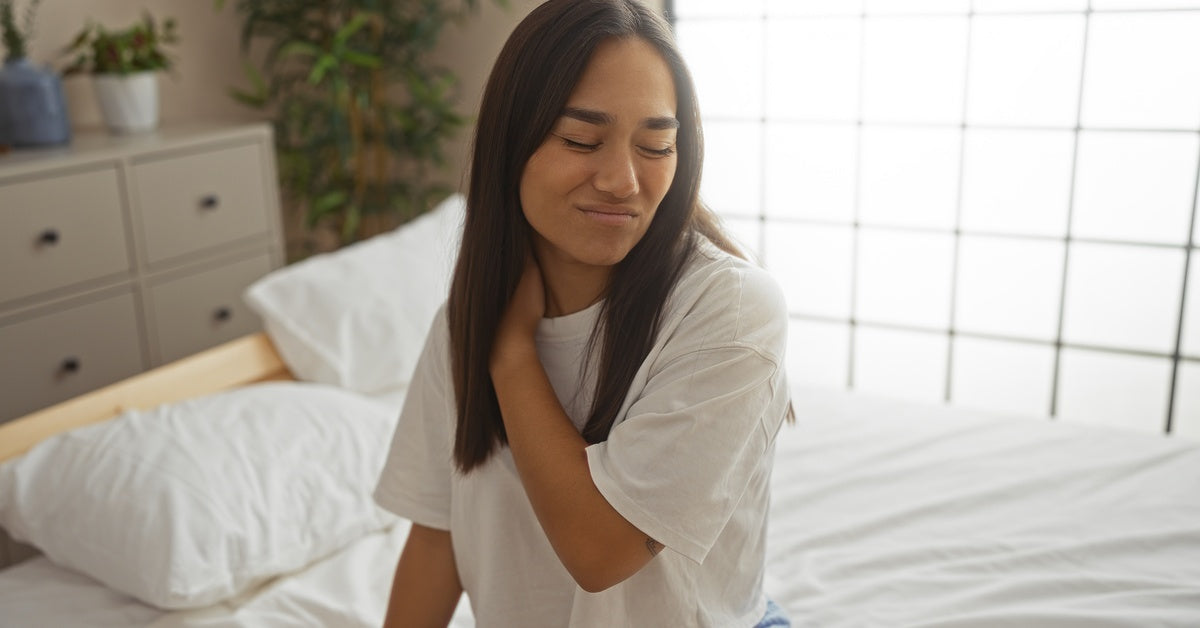 A woman sits on a clean and white bed and makes a pained face while she massages her right shoulder and neck.